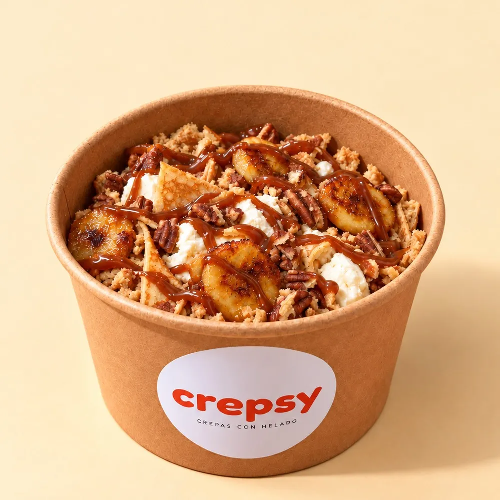 Bowl de papel con crepa y helado, plátano dorado, nuez pecana, galleta molida y cajeta en hilos
